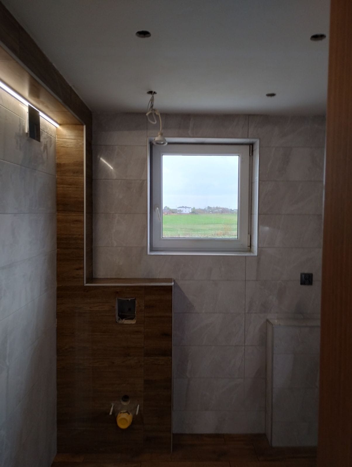 Łazienka w trakcie wykańczania: marmurowe płytki ścienne, drewnopodobne akcenty, okno z widokiem na zielone pole. Widoczne niedokończone instalacje.