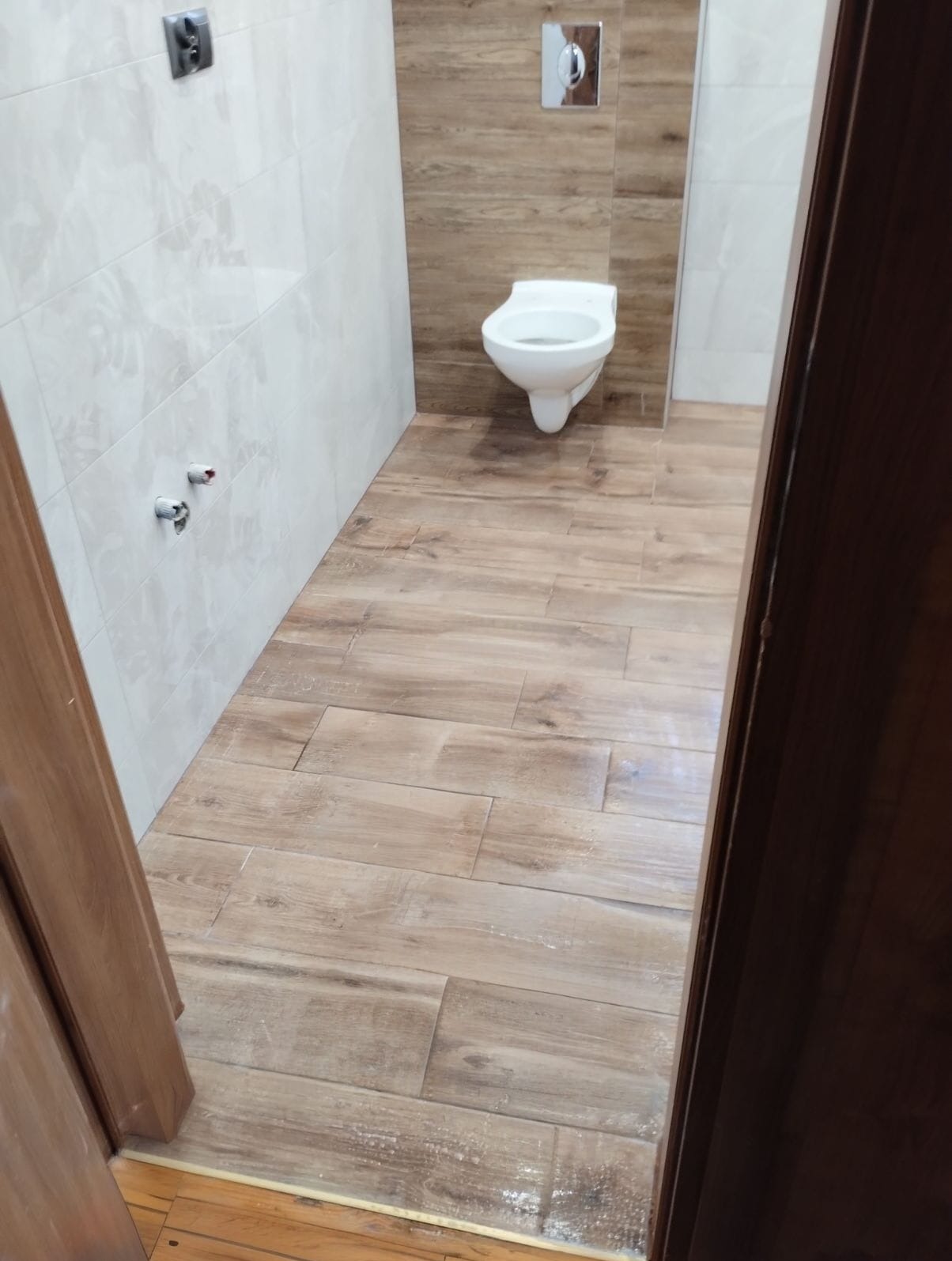 Ukończona łazienka z toaletą i płytkami imitującymi drewno na podłodze i ścianie. Widok zza otwartych, drewnianych drzwi. Jasne płytki na ścianach z lewej.
