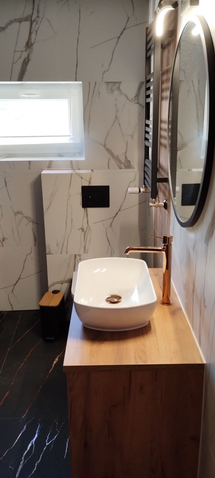 Nowoczesna łazienka z umywalką nablatową, baterią w kolorze różowego złota i marmurowymi płytkami. Minimalistyczny styl, czarne akcenty i okrągłe lustro.