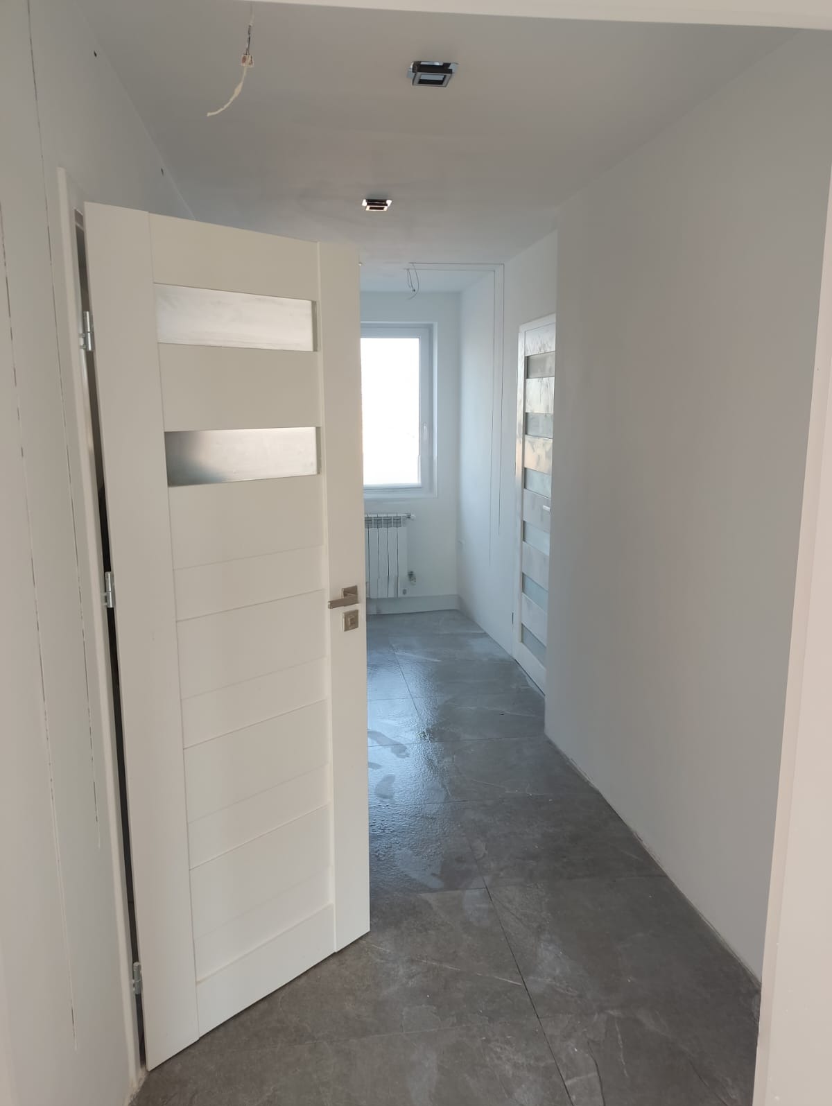 Białe, częściowo otwarte drzwi z przeszkleniami w nowym wnętrzu. Szare płytki podłogowe, świeżo pomalowane ściany i widok na okno w oddali. Minimalistyczny design.