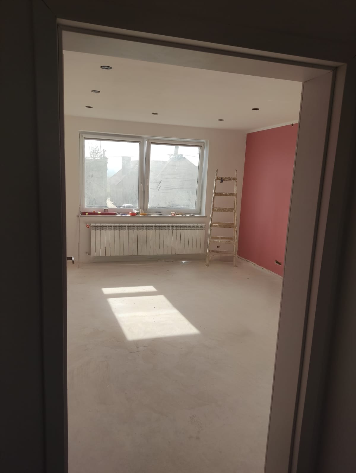 Pokój po szpachlowaniu, przygotowany do malowania. Widoczne gładkie ściany, okno z grzejnikiem i drabina malarska. Świeżo wykończone wnętrze z różową ścianą.