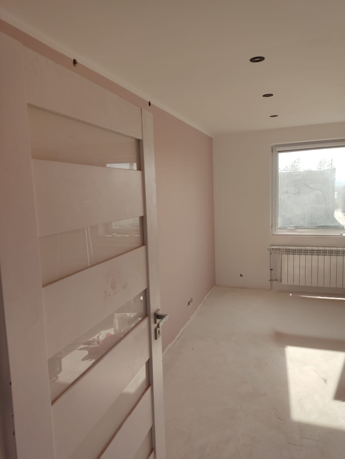 Pokój po remoncie: ściany w odcieniach różu i bieli, białe drzwi z przeszkleniami, grzejnik pod oknem. Widoczne ślady po pracach na podłodze.