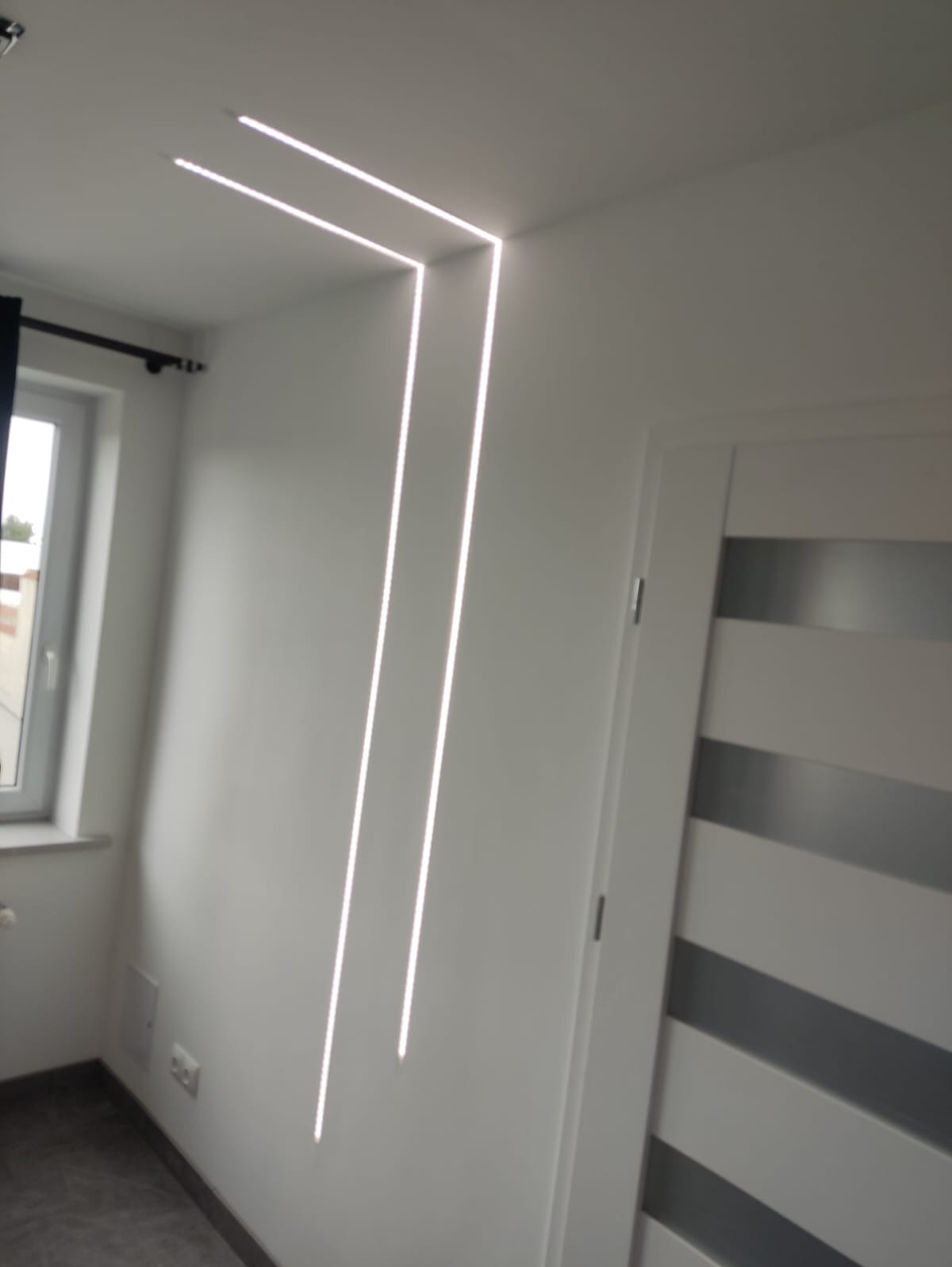 Białe wnętrze z oknem i drzwiami. Na suficie i ścianie zamontowane nowoczesne, liniowe oświetlenie LED, tworzące geometryczny wzór. Minimalistyczny design.