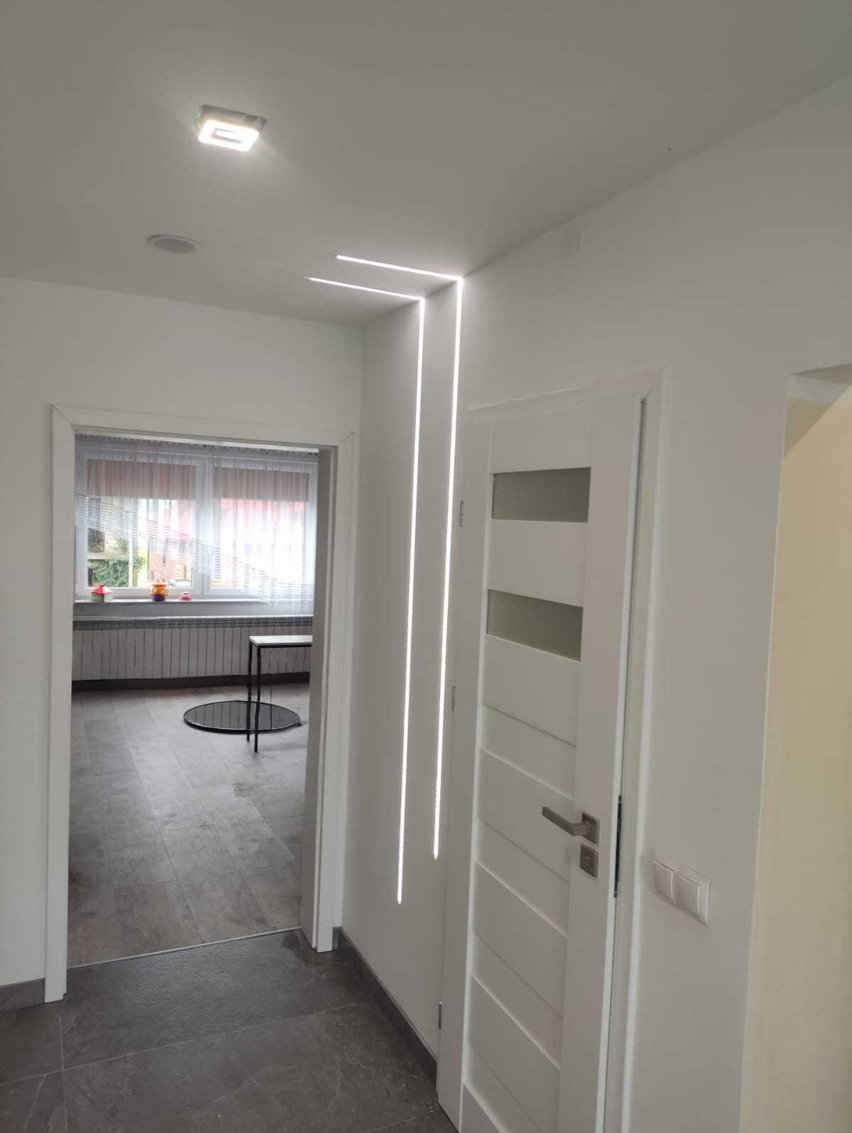 Białe wnętrze z nowoczesnym oświetleniem LED wzdłuż ściany, widok na pokój z szarymi płytkami i białą stolarką okienną. Białe drzwi wewnętrzne z przeszkleniami.
