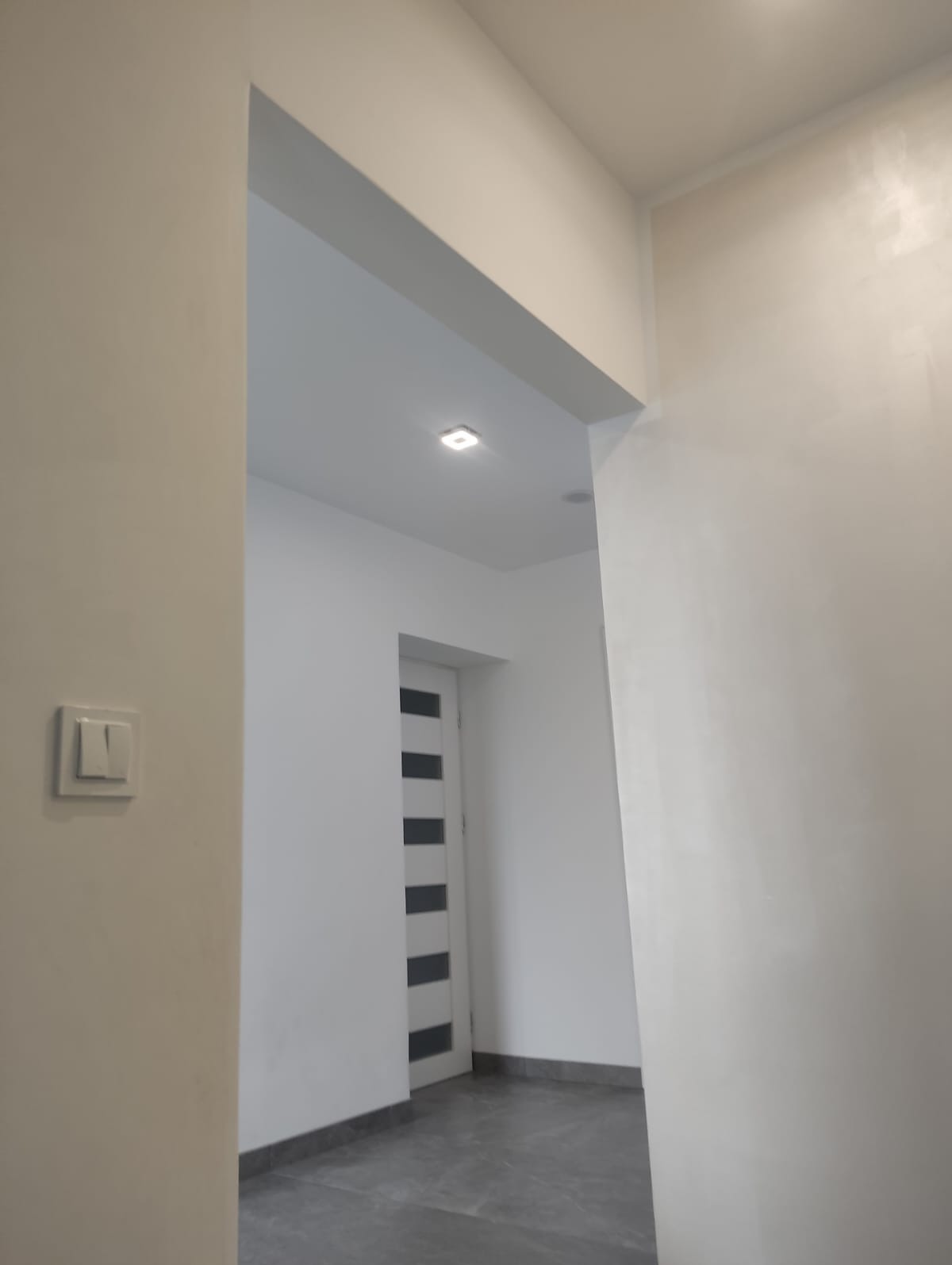 Białe, świeżo pomalowane wnętrze z widocznym włącznikiem światła, fragmentem sufitu z oświetleniem punktowym i częściowo widocznymi drzwiami z przeszkleniami.