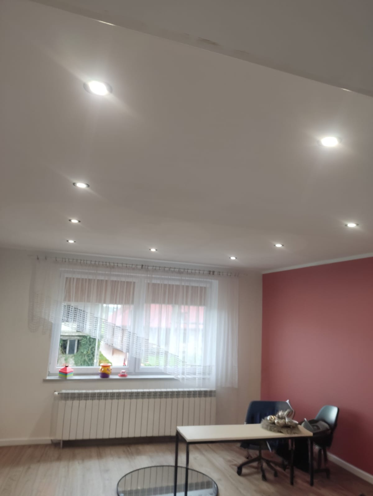 Jasne wnętrze pokoju po remoncie: biały sufit z punktowym oświetleniem LED, okno z firanką, ściana w odcieniu różu i drewniana podłoga. Widoczny grzejnik i meble.