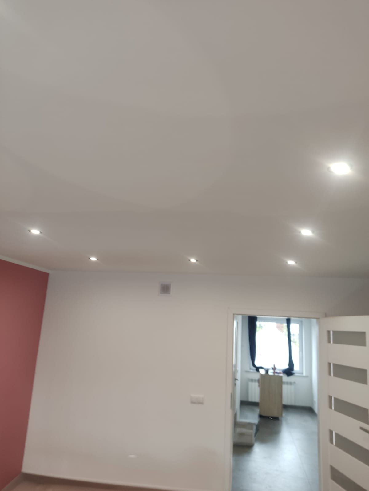 Wnętrze po malowaniu: białe ściany z akcentem bordowej ściany, sufit z oświetleniem punktowym, widok na pokój przez otwarte drzwi z białymi panelami.