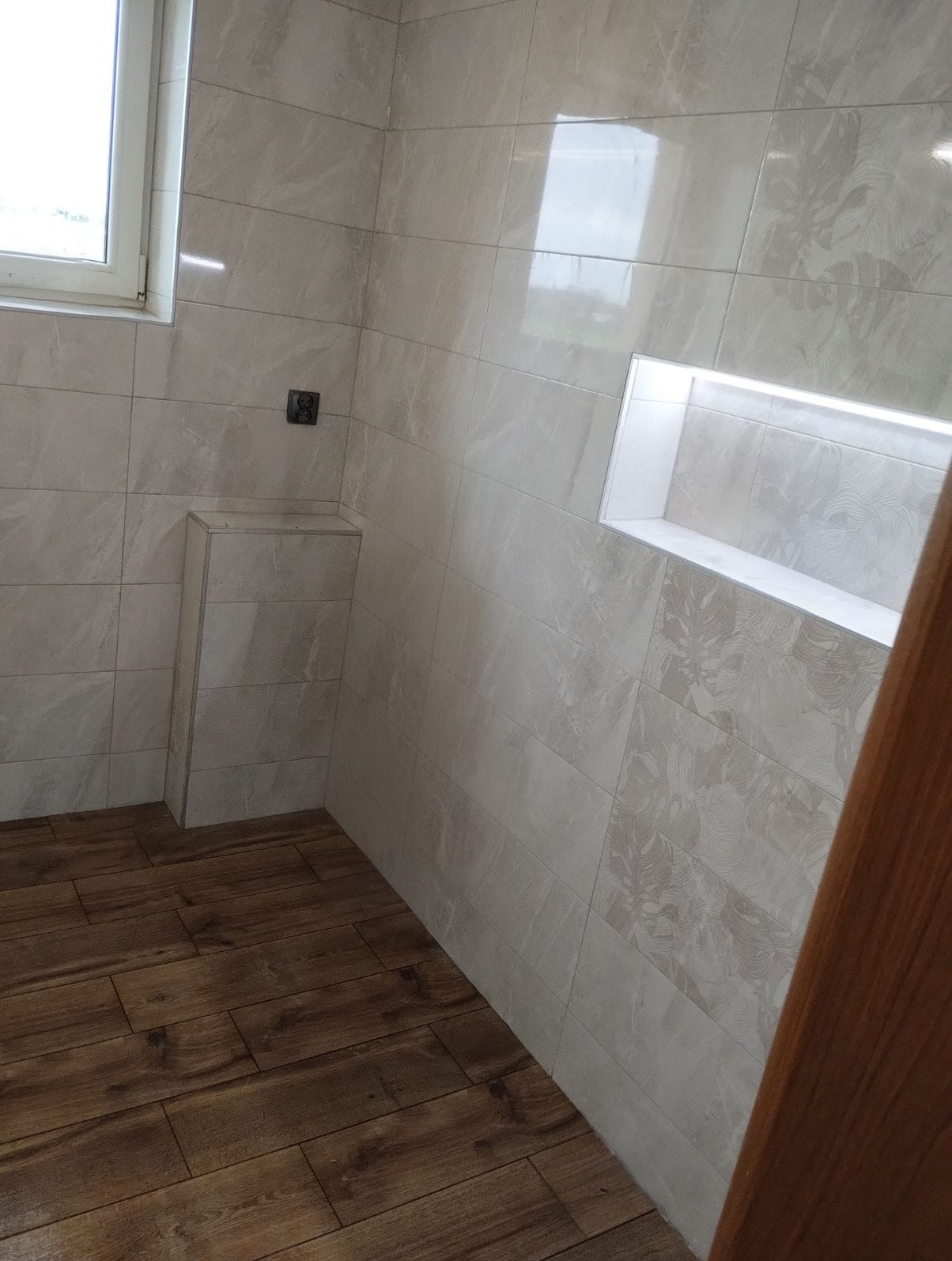 Jasna łazienka po remoncie: drewnopodobna podłoga, płytki ścienne z subtelnym wzorem, wnęka z oświetleniem LED i zabudowa z płytek. Widok na fragment okna.