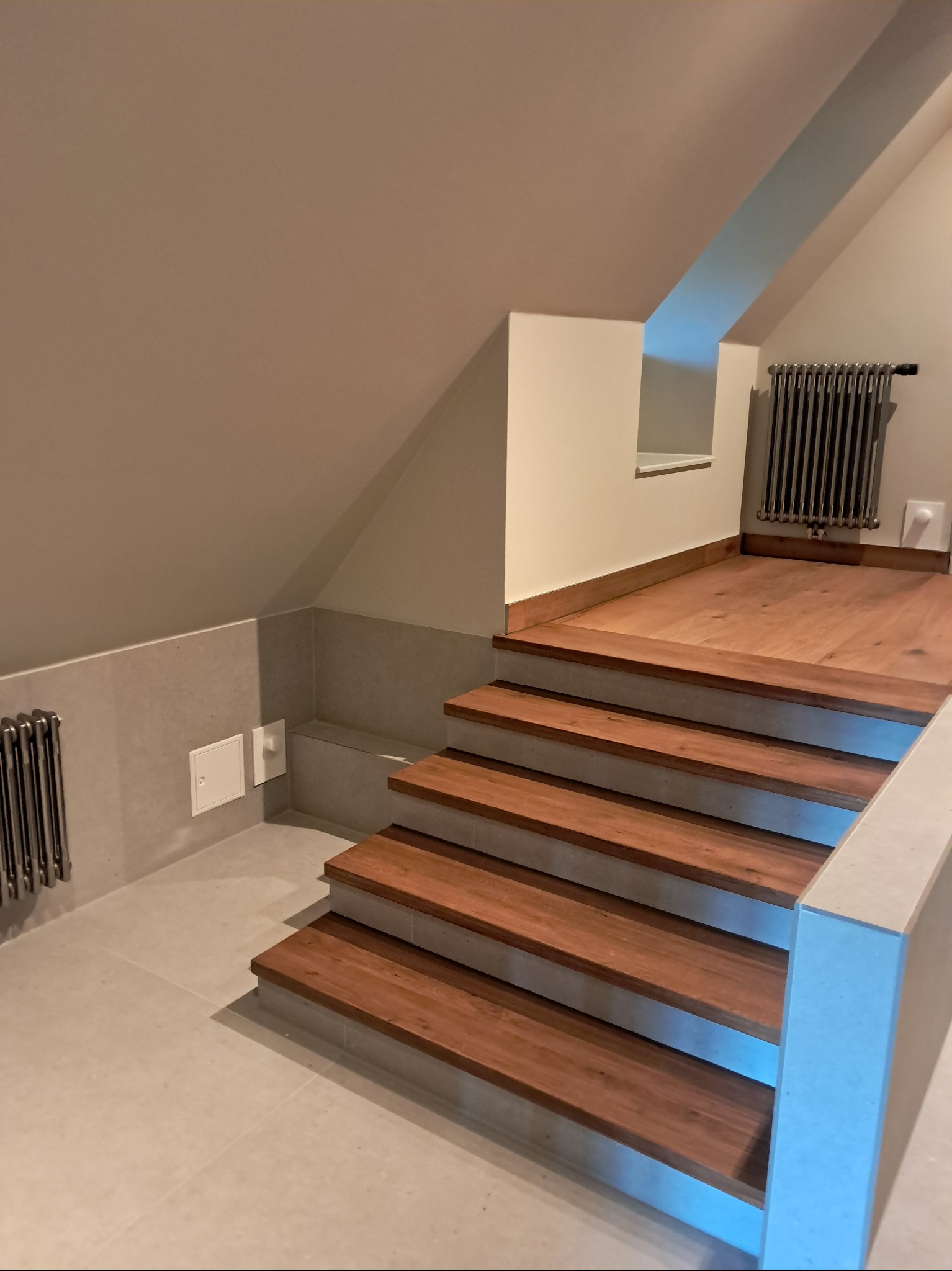 Drewniane schody z szarymi elementami w jasnym wnętrzu poddasza. Grzejnik żeberkowy na ścianie. Ujęcie z dołu, widoczne stopnie i fragment podłogi.