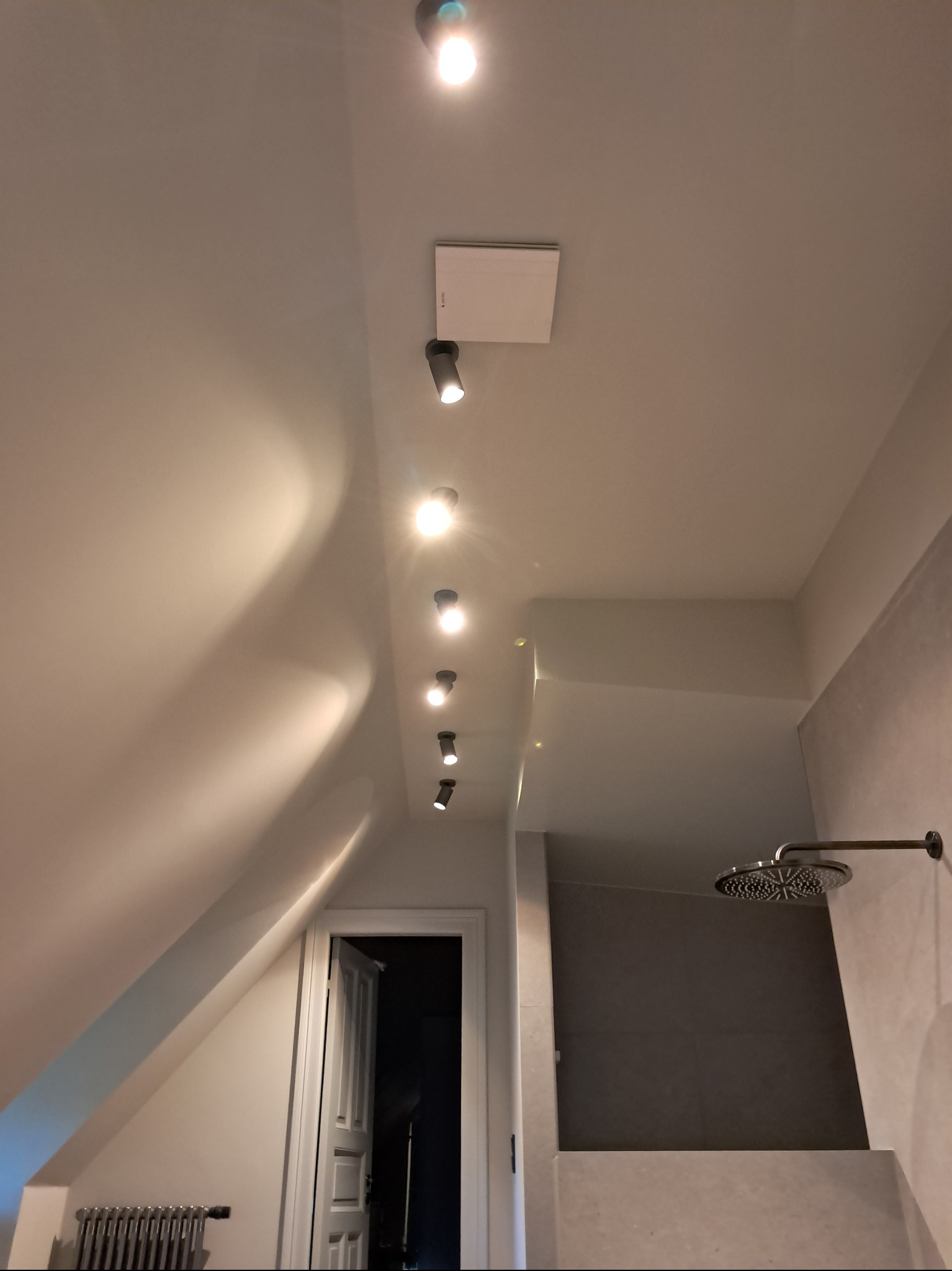 Nowoczesna łazienka na poddaszu z oświetleniem punktowym, szarą kabiną prysznicową i grzejnikiem żeberkowym. Ujęcie z dołu, podkreślające skosy i minimalistyczny design.