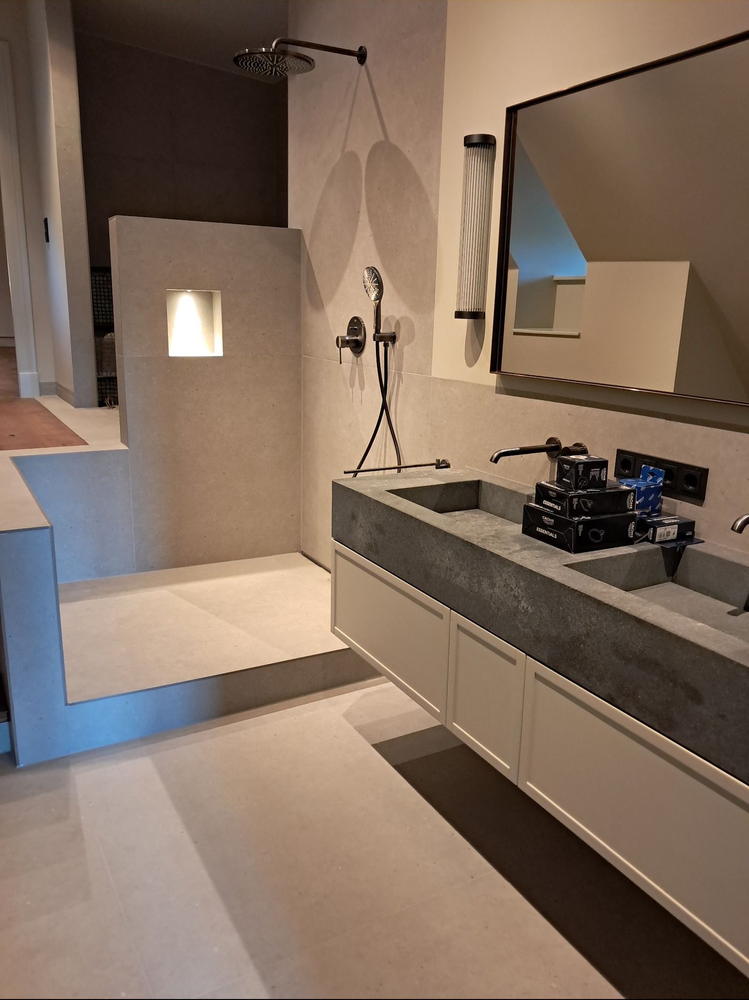 Nowoczesna łazienka z kabiną prysznicową typu walk-in, umywalką z szarego kamienia i minimalistycznym oświetleniem. Na blacie umywalki pudełka z akcesoriami.