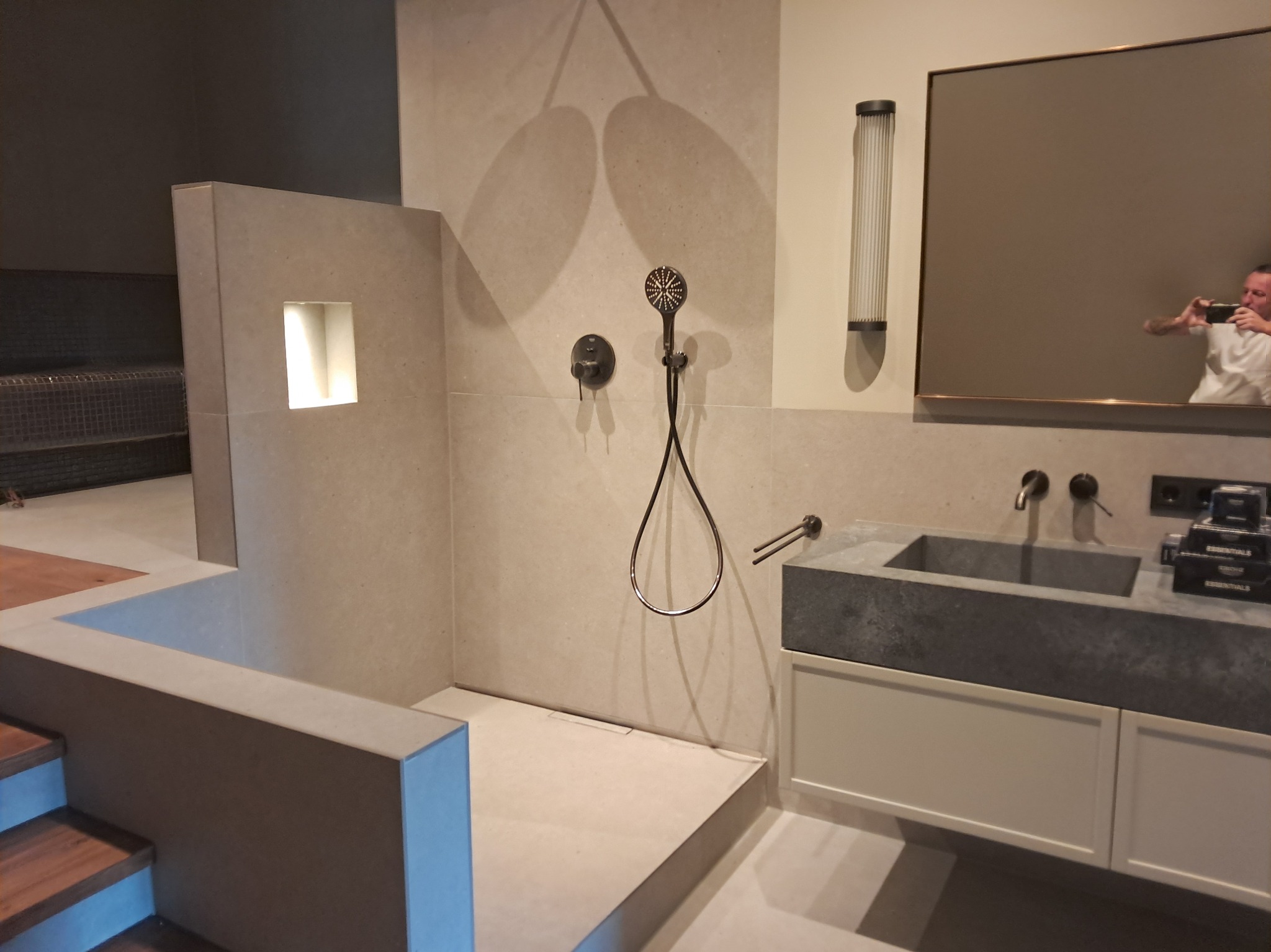 Nowoczesna łazienka z kabiną prysznicową typu walk-in, umywalką z szarego kamienia i dużym lustrem, w którym odbija się osoba robiąca zdjęcie.