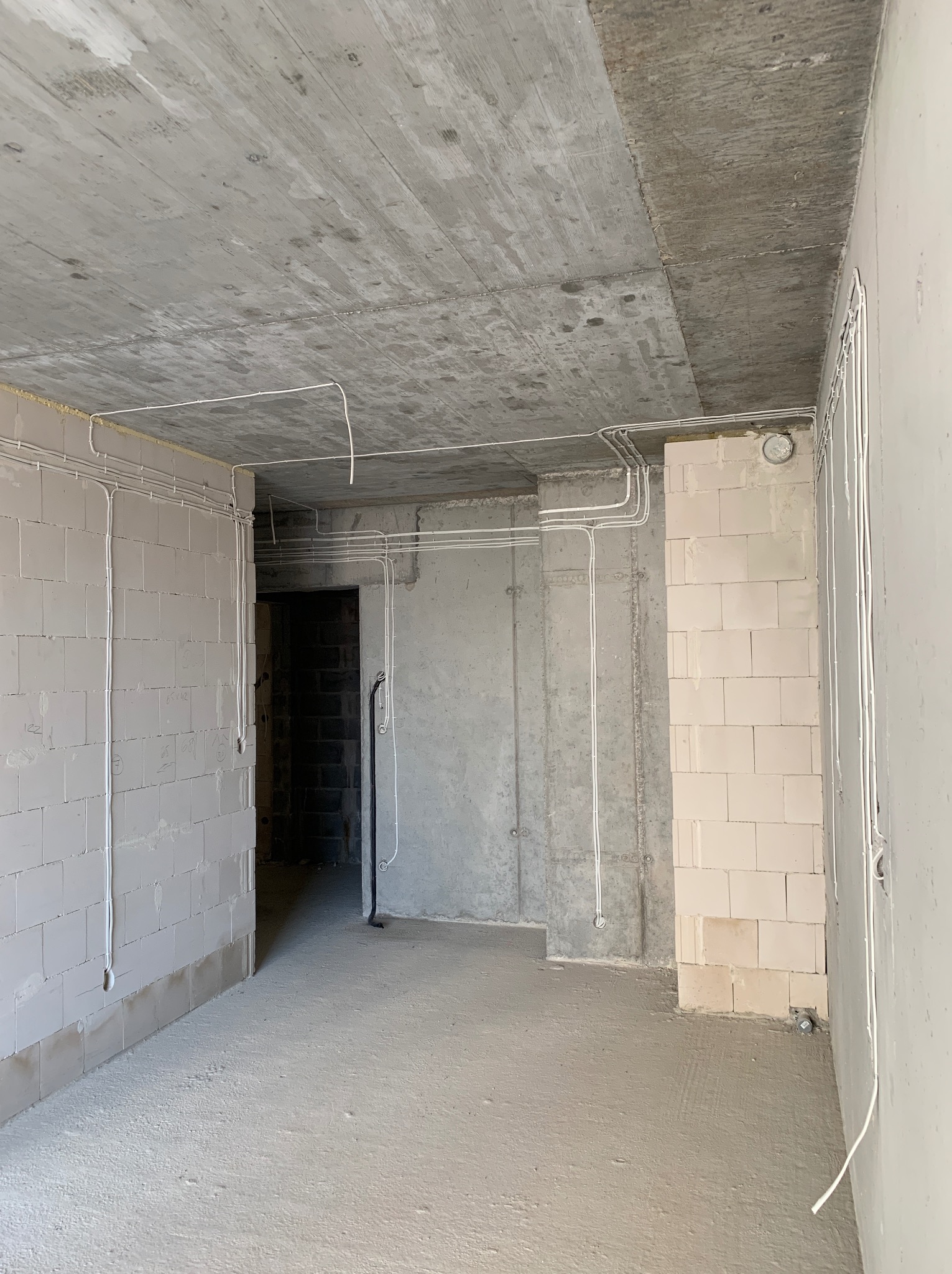 Surowy pokój w trakcie prac wykończeniowych z widocznymi przewodami elektrycznymi poprowadzonymi po ścianach i suficie, ściany z betonu i bloczków, widoczny otwór drzwiowy.