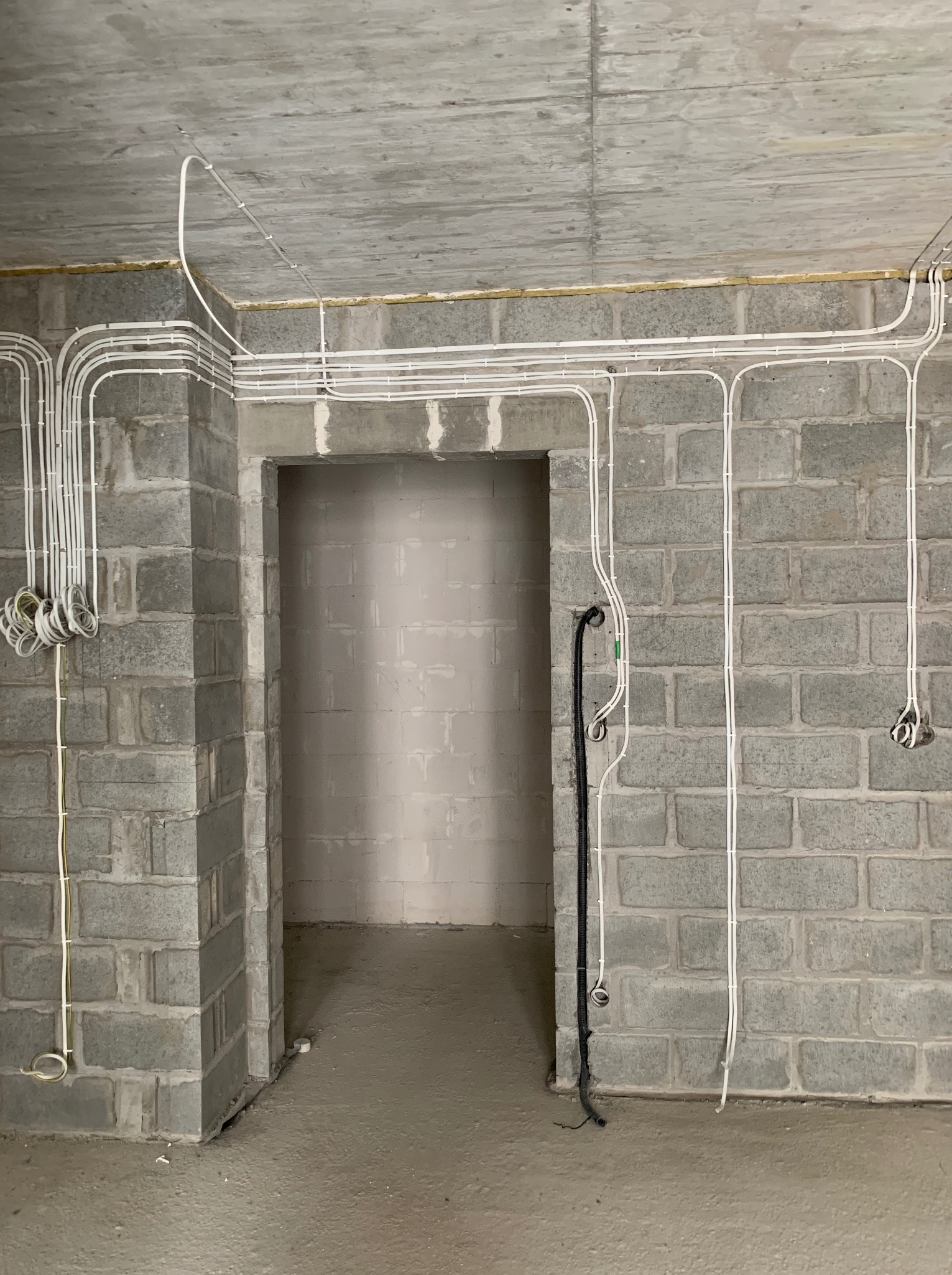 Surowe wnętrze w trakcie prac: widoczne szare ściany z betonu komórkowego, surowy betonowy sufit i liczne białe kable elektryczne prowadzone po powierzchni ścian i sufitu, niektóre zakończone...