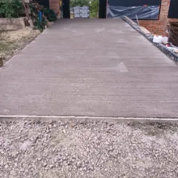 Świeżo wylana, gładka, betonowa posadzka przemysłowa prowadząca do budynku gospodarczego z zapasem materiałów budowlanych w tle. Widok z dołu, na pierwszym planie żwir.