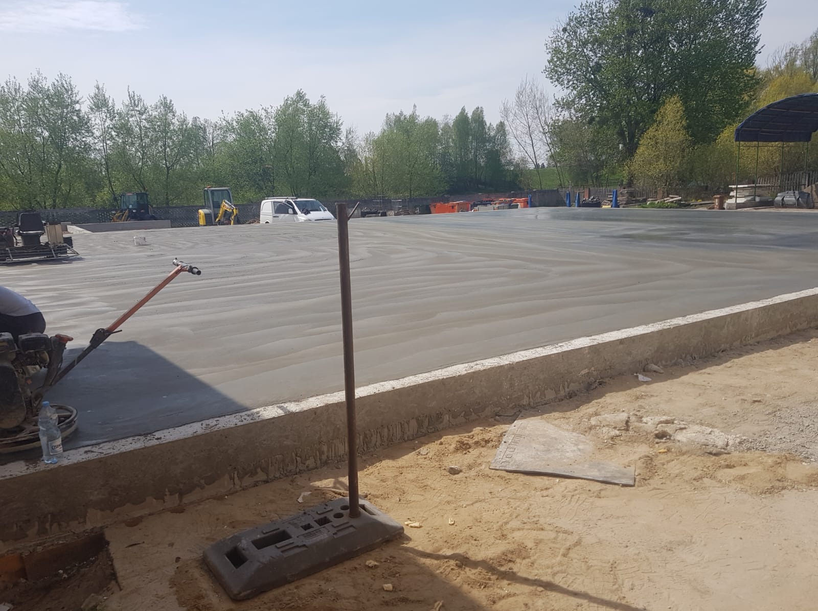 Świeżo wylana, gładka posadzka betonowa na zewnątrz budynku, wyrównywana maszynowo. W tle drzewa, maszyny budowlane i biały samochód dostawczy.