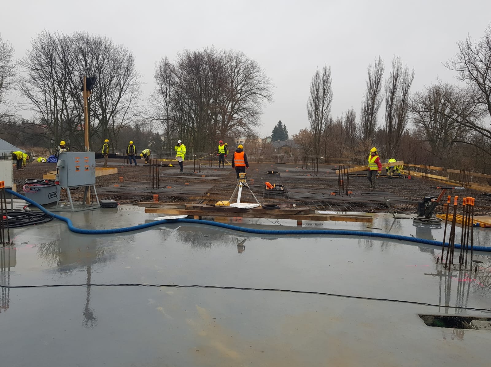 Wylewka samopoziomująca w trakcie realizacji. Widoczne zbrojenia, ekipa w strojach roboczych i sprzęt. W tle drzewa i szara pogoda.