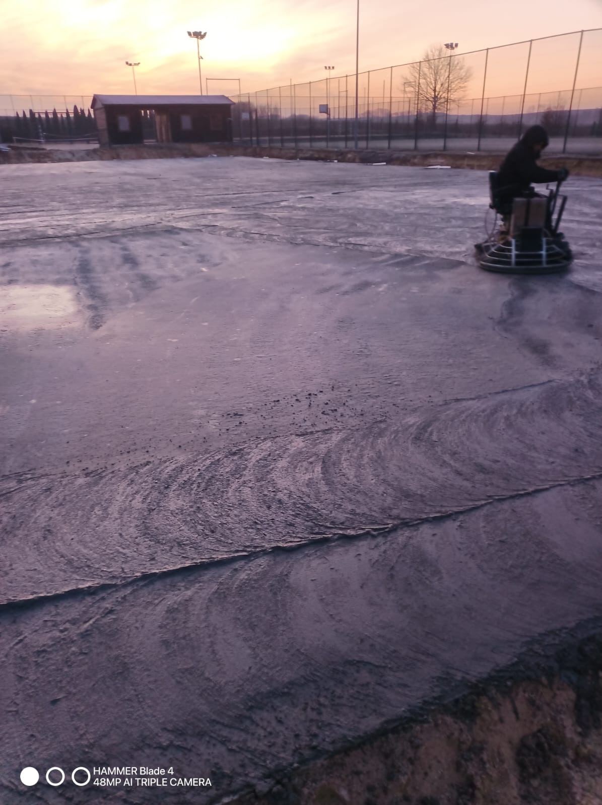 Praca na zewnątrz: operator maszyny wygładza świeżo wylaną betonową powierzchnię na tle zachodzącego słońca i budynku w oddali.