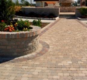 Ułożona nawierzchnia z beżowej kostki brukowej z bordurowym obramowaniem, prowadząca do schodów. Z lewej strony okrągła rabata kwiatowa z cegły, a w tle widoczny budynek.