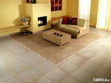 Jasne płytki podłogowe w salonie z kominkiem i beżową sofą. W centralnej części wzór z płytek w odcieniach brązu, tworzący dywan. Ściany w odcieniach żółci, nad kominkiem obraz.