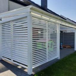 Biała, nowoczesna konstrukcja z poziomych listew, przylegająca do domu. Widoczne oświetlenie LED pod dachem, kostka brukowa i trawnik. Słoneczny dzień.