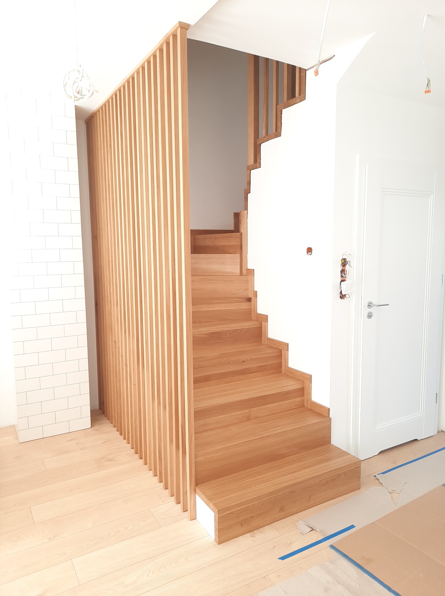 Jasne drewniane schody z ażurową balustradą z pionowych listew, widoczne zabezpieczenia podłogi. Minimalistyczny design, naturalne światło, nowoczesne wykończenie wnętrza.