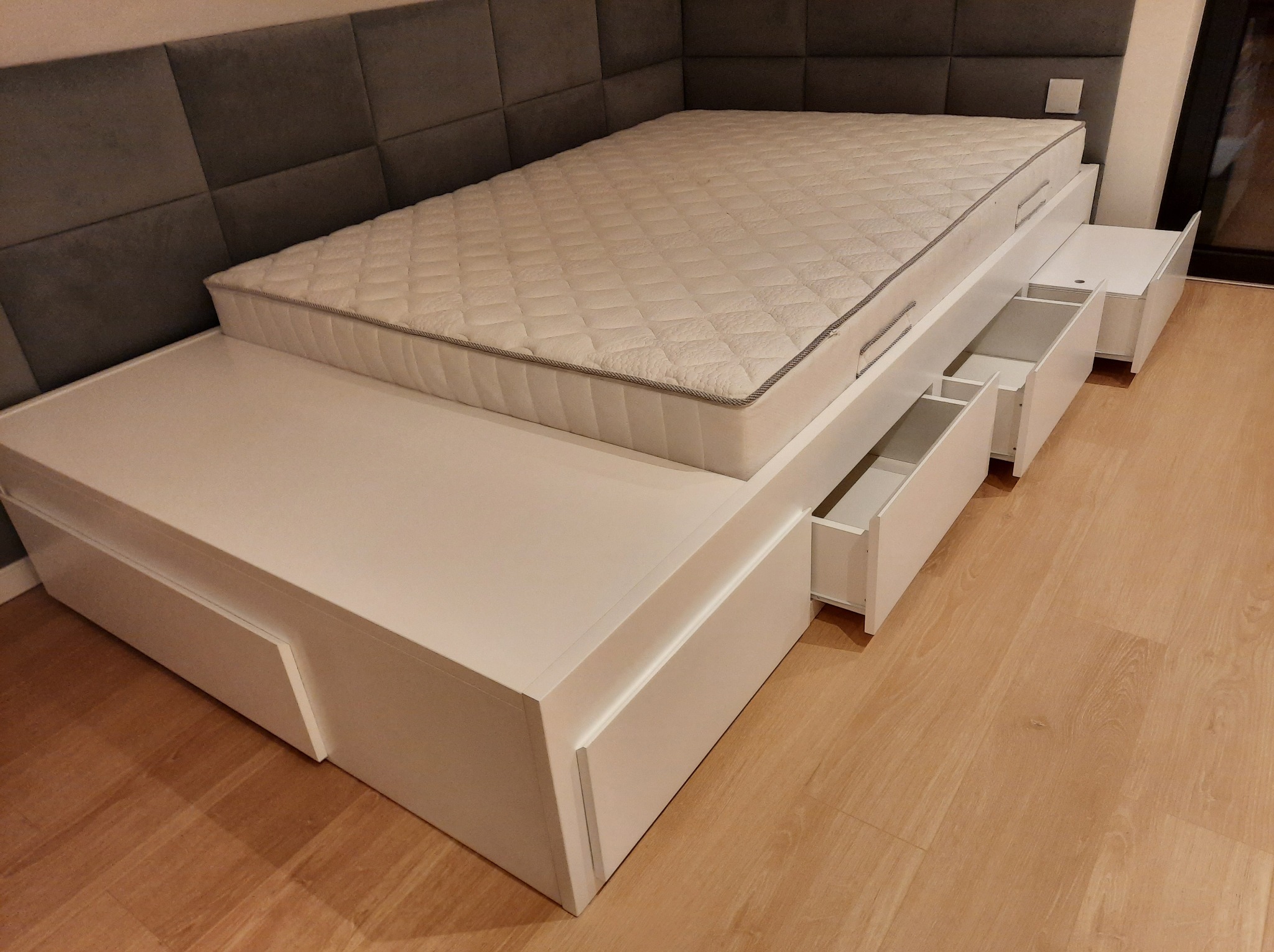 Białe łóżko z szufladami i materacem na drewnianej podłodze. Częściowo otwarte szuflady ukazują wnętrze. Szary zagłówek w panelach. Ujęcie z niskiego kąta.