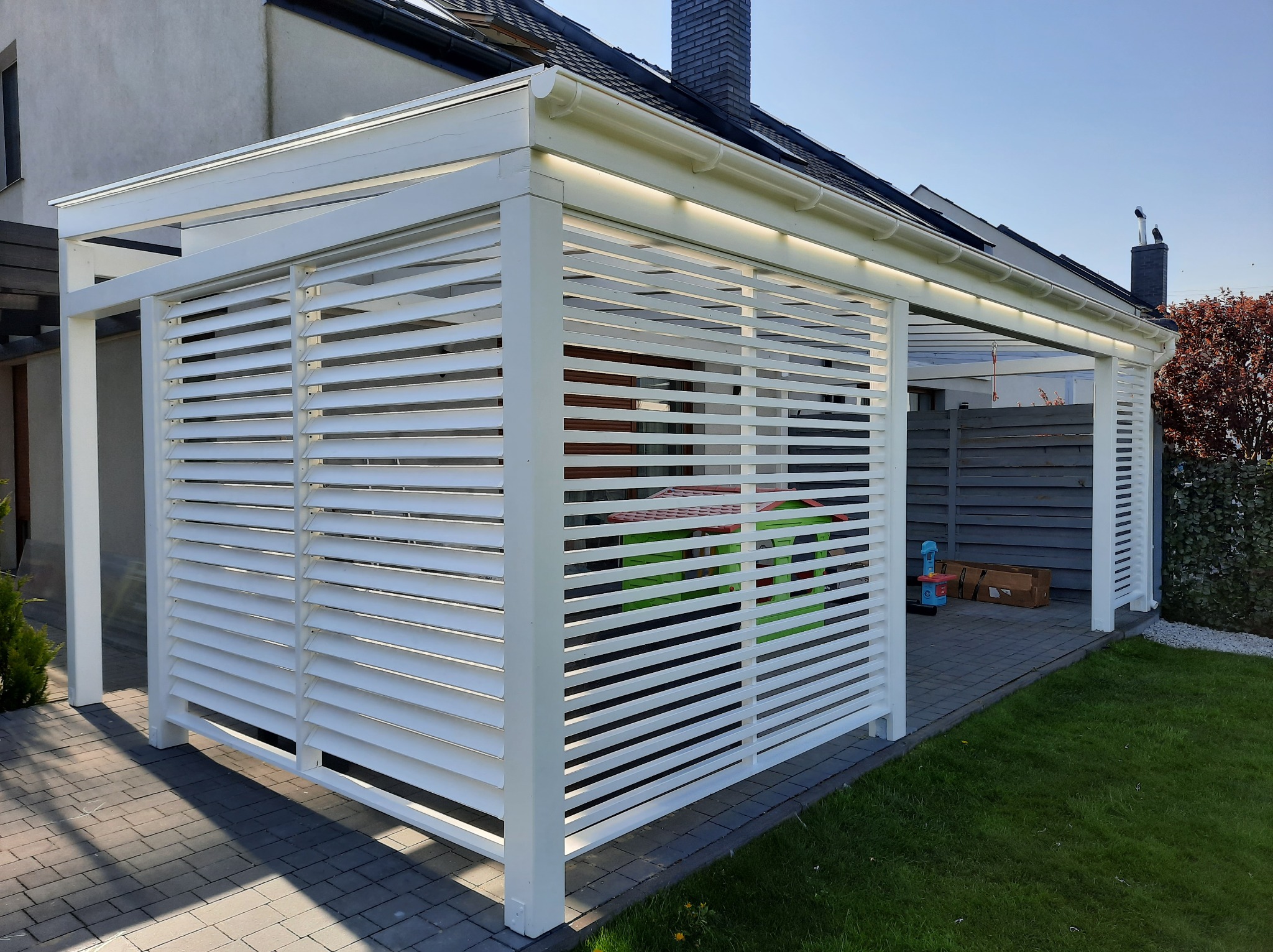 Biała, nowoczesna konstrukcja z poziomych listew, przylegająca do domu. Widoczne oświetlenie LED pod dachem, kostka brukowa i trawnik. Słoneczny dzień.