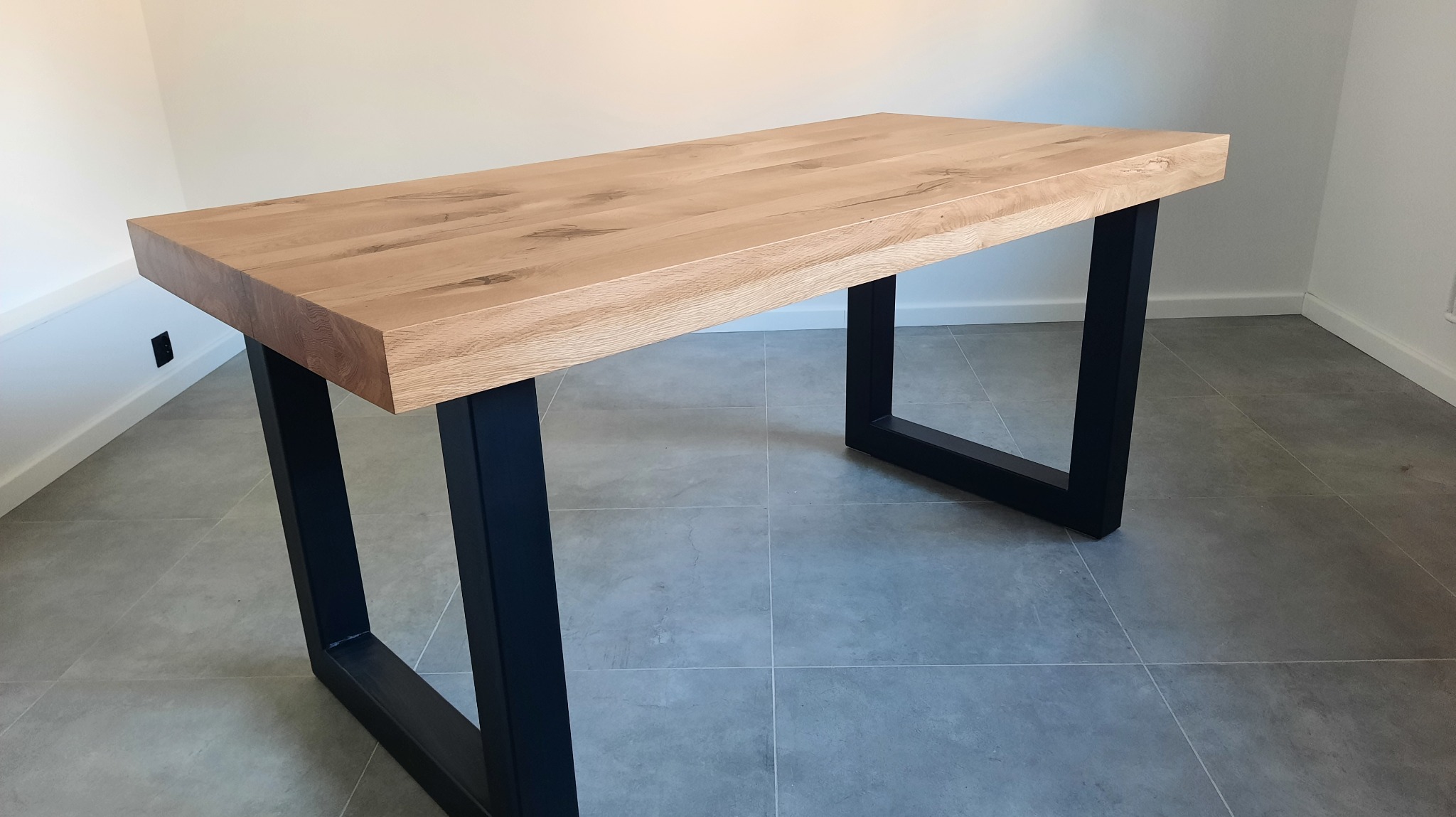 Stół z jasnego drewna o grubym blacie i czarnych, metalowych nogach w kształcie prostokątów. Minimalistyczny design na tle szarej podłogi z płytek.