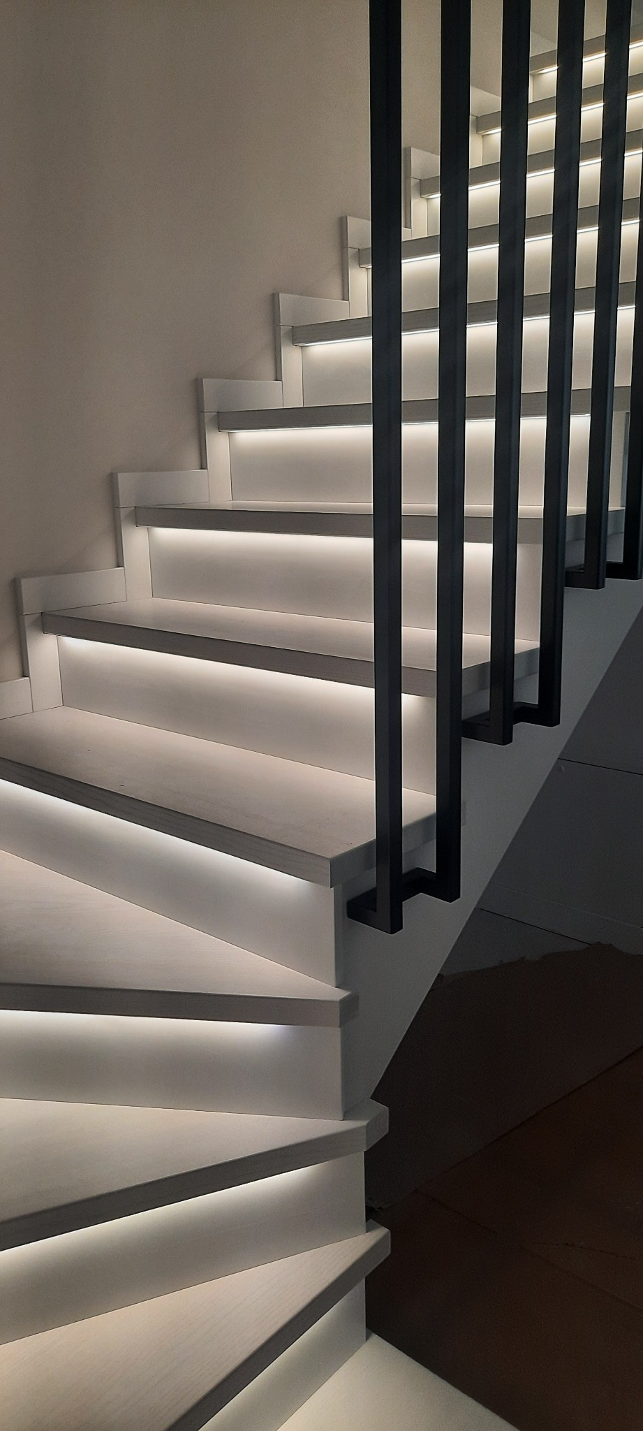 Nowoczesne schody z podświetleniem LED pod stopniami i czarną, minimalistyczną balustradą. Neutralna kolorystyka, ciepłe światło. Widok z dołu.