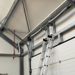 P&O WORKS SPÓŁKA Z OGRANICZONĄ ODPOWIEDZIALNOŚCIĄ - Wnętrze hali z metalową konstrukcją dachu i ścianami z paneli. Aluminiowa drabina oparta o ścianę. Widoczne elementy instalacji pod sufitem.