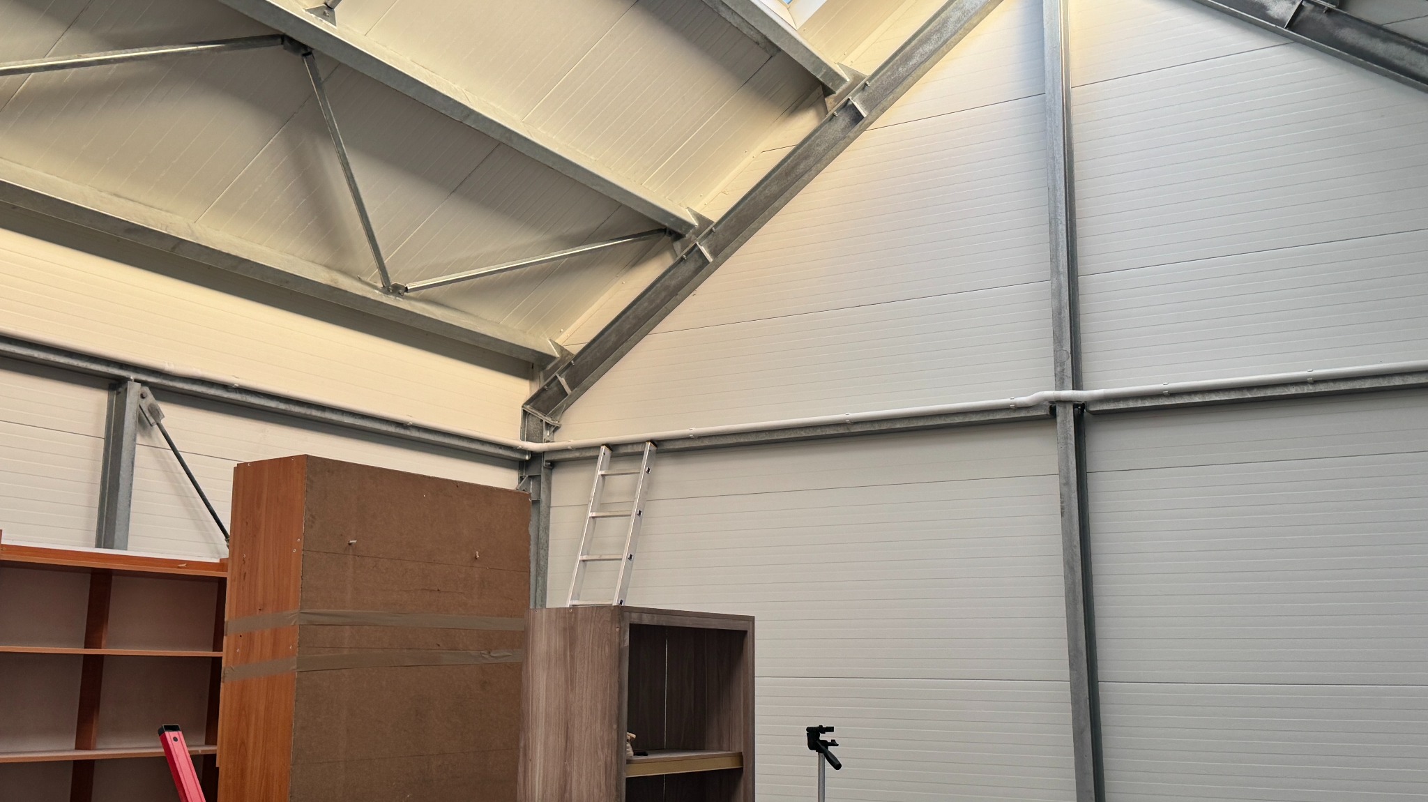 Wnętrze hali z widoczną konstrukcją dachu, ścianami z paneli, drabiną i meblami w trakcie montażu. Widoczne elementy instalacji pod sufitem.