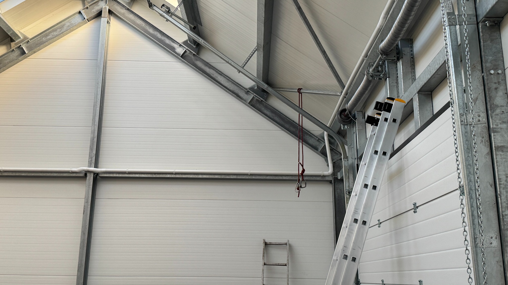 Wnętrze hali z metalową konstrukcją dachu i ścianami z paneli. Widoczna drabina, rury wentylacyjne i elementy mocujące. Oświetlenie górne, surowe wykończenie.