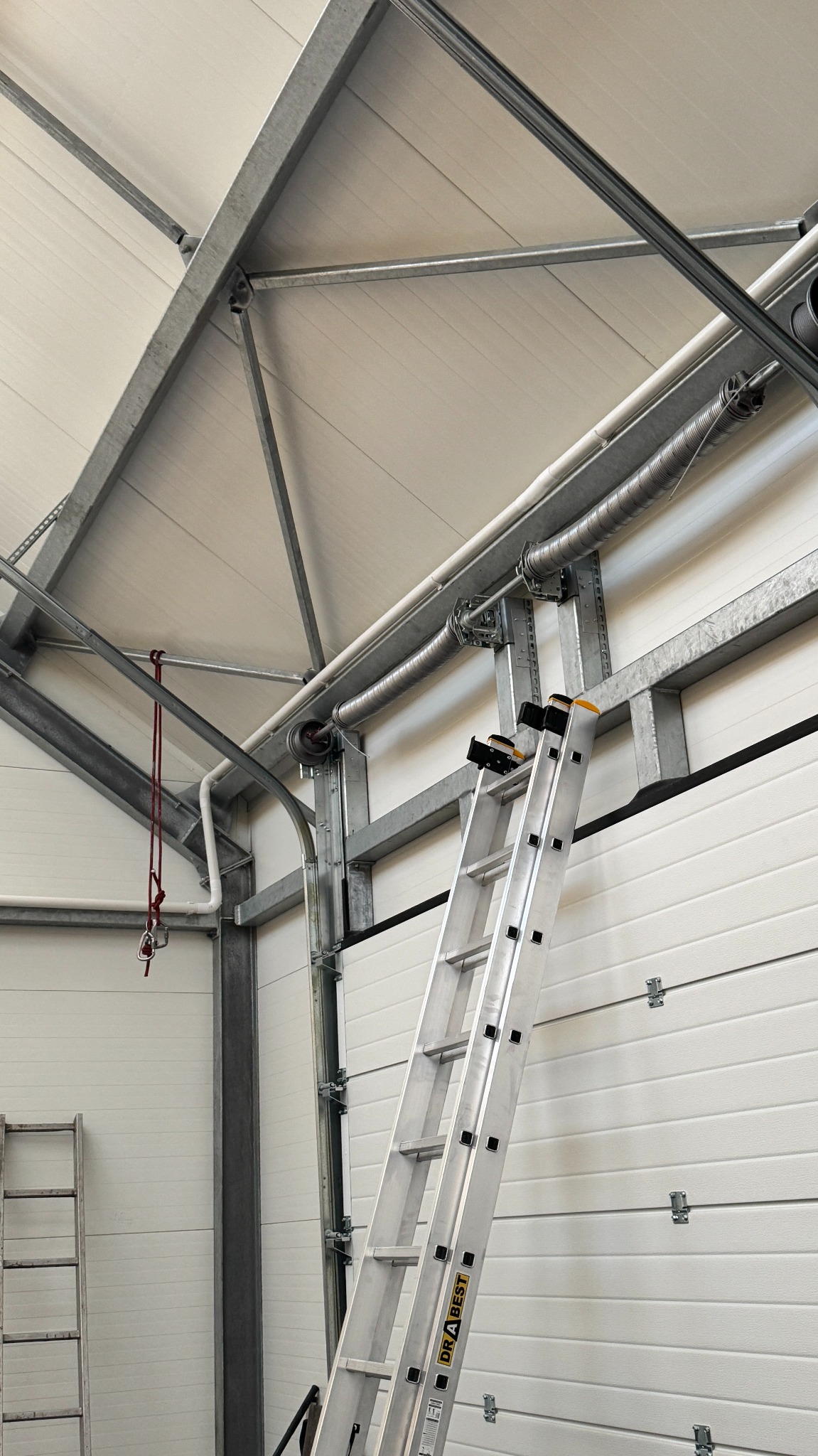 Wnętrze hali z metalową konstrukcją dachu i ścianami z paneli. Aluminiowa drabina oparta o ścianę. Widoczne elementy instalacji pod sufitem.