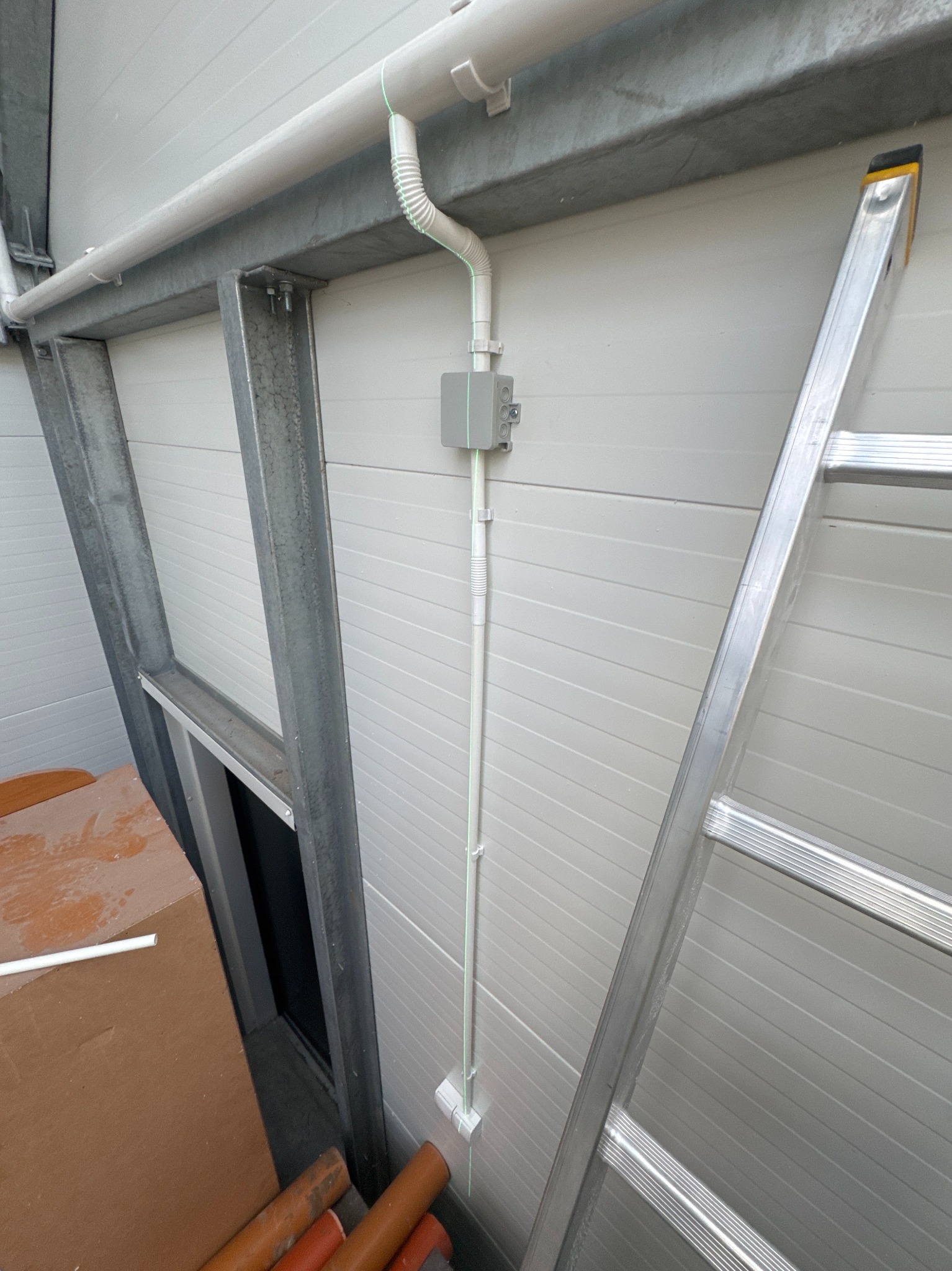 Szara skrzynka elektryczna z rurką na ścianie z blachy trapezowej, obok drabina aluminiowa. Widoczne rury i elementy konstrukcyjne budynku.