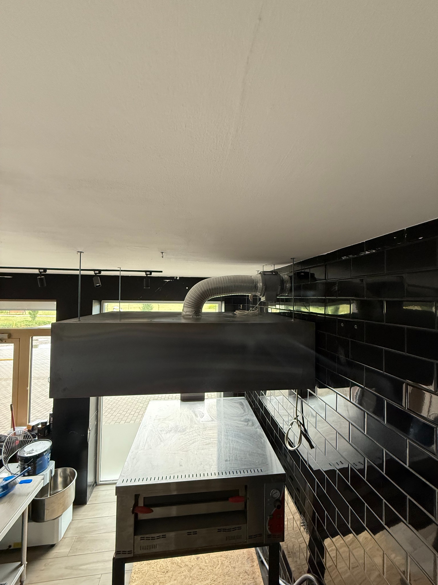 Zamontowany okap kuchenny ze stalowym przewodem wentylacyjnym w lokalu gastronomicznym, widok z dołu, czarne płytki na ścianie, oświetlenie punktowe.