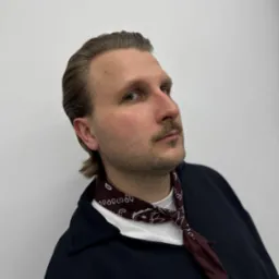 Mężczyzna z zaczesanymi do tyłu włosami, wąsami i bordową apaszką pod szyją, spoglądający w górę po zabiegu stylizacji.