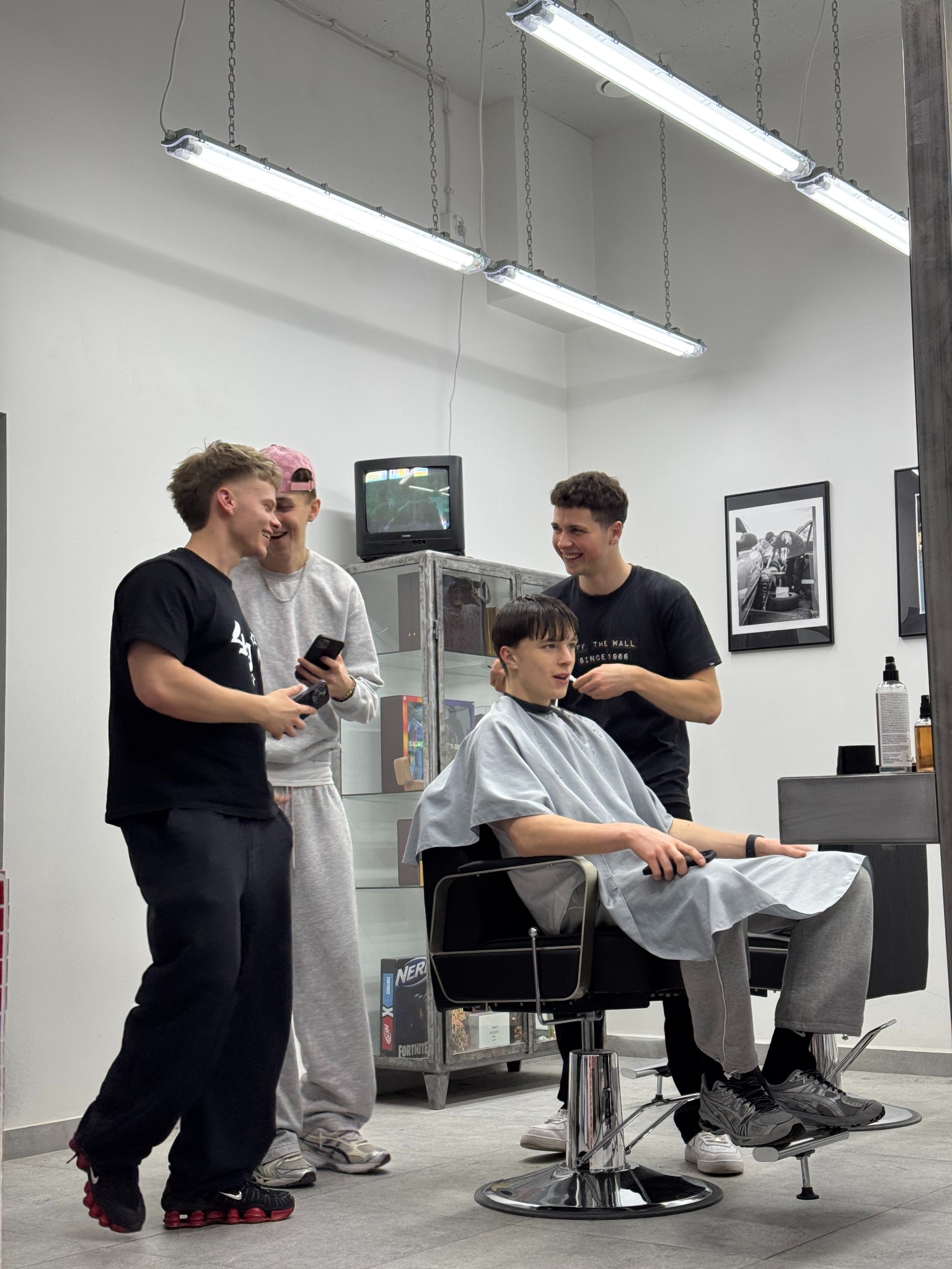 Młody mężczyzna w pelerynie fryzjerskiej siedzi na fotelu, uśmiechnięty, podczas gdy fryzjer poprawia mu włosy, w tle dwóch kolegów obserwuje i robi zdjęcia telefonem w jasnym salonie.