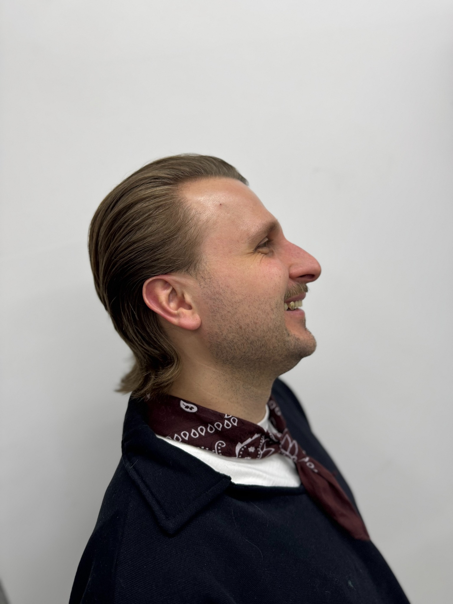 Mężczyzna z zaczesanymi do tyłu włosami, w profilu, uśmiechnięty, z chustą na szyi