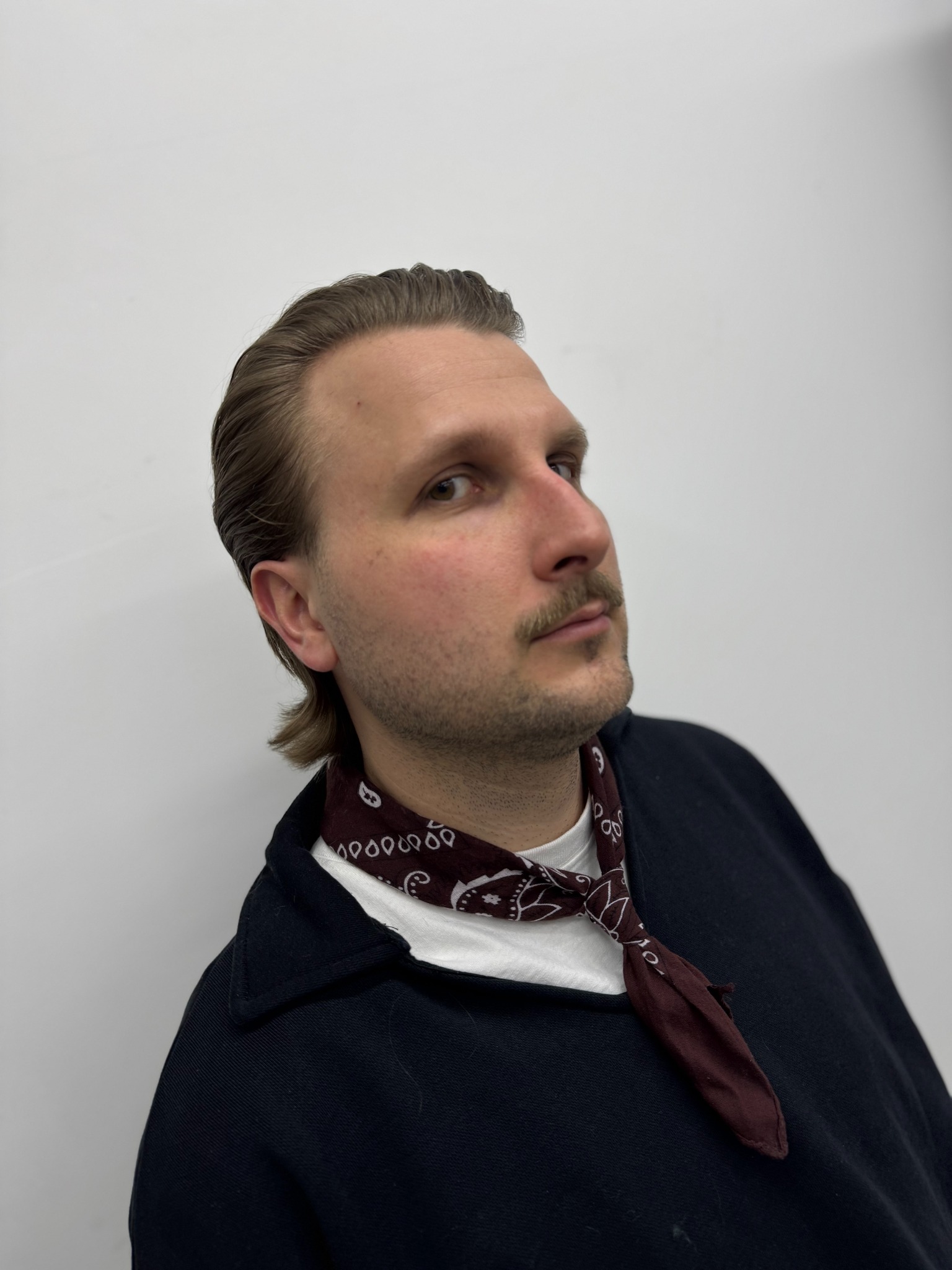 Mężczyzna z zaczesanymi do tyłu włosami, wąsami i bordową apaszką pod szyją, spoglądający w górę po zabiegu stylizacji.