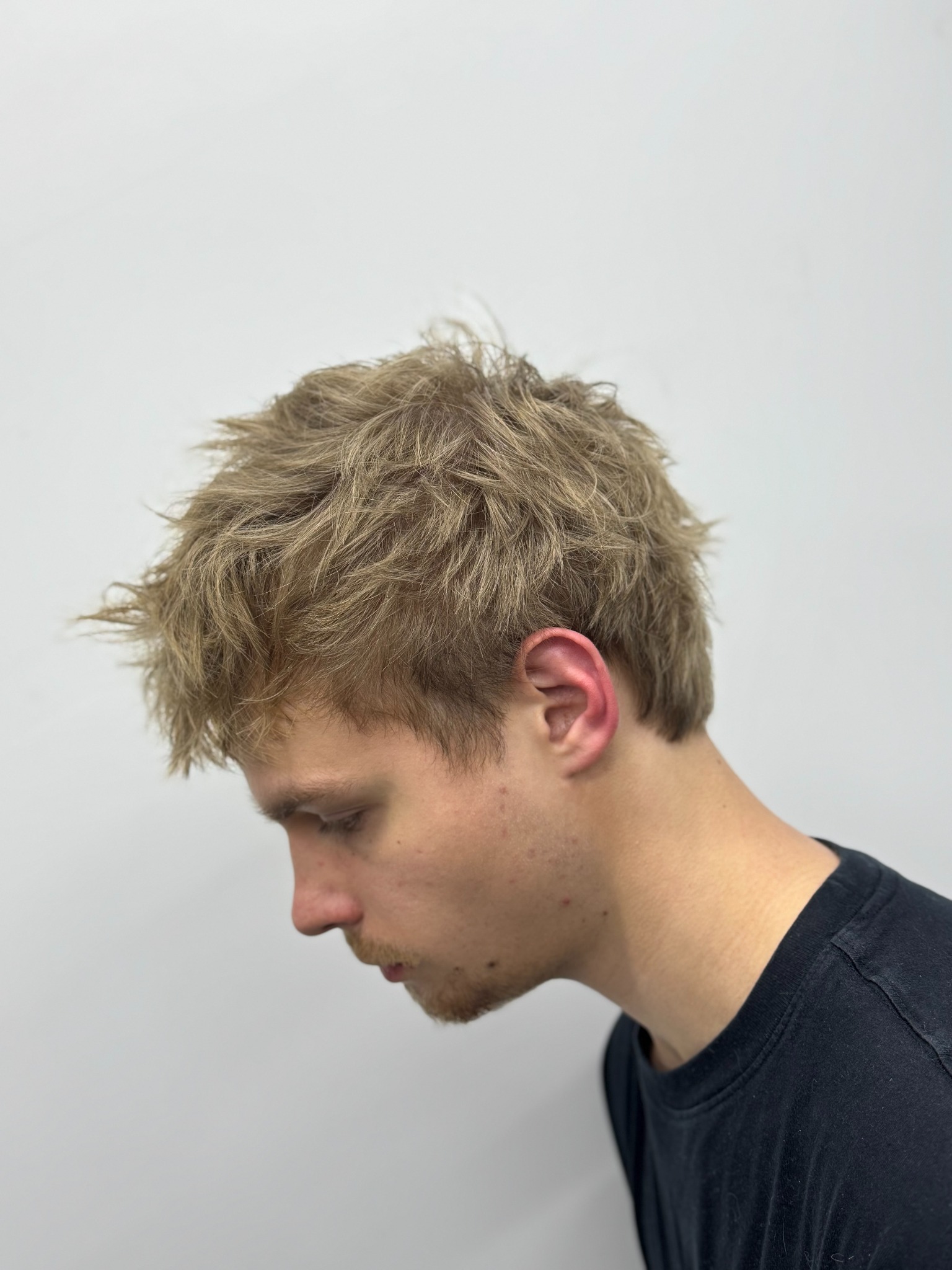 Młody mężczyzna z blond, potarganą fryzurą, profil głowy w dół na tle białej ściany.