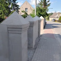 Świeżo otynkowane słupki ogrodzeniowe z piramidalnymi zwieńczeniami, widoczne z boku na tle ulicy i domów. Detal wykończenia zewnętrznego w jasnym świetle.