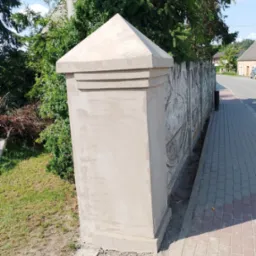 Wykończony fragment ogrodzenia z gładzią i dekoracyjnym wzorem, obok chodnik z kostki. Widoczne krzewy i drzewa w tle. Słoneczny dzień.