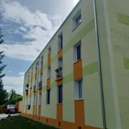 Odnowiona elewacja bloku z balkonami, pomalowana w poziome pasy: żółto-zielone i pomarańczowe. Widoczne okna, rynny i fragment trawnika. Słoneczny dzień.