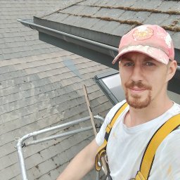 Tomasz Sitarz - Pracownik na rusztowaniu, w uprzęży, na tle dachu pokrytego dachówką z mchem, w trakcie pracy. Widoczny fragment rynny i okno dachowe.