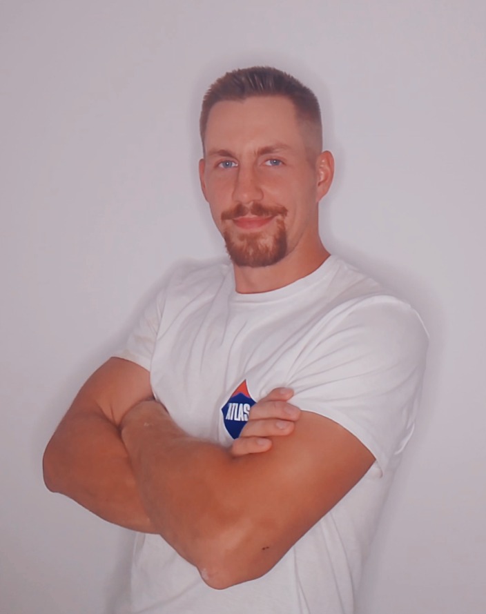 Młody mężczyzna z brodą, w białej koszulce z logo Atlas, stoi na tle jasnej ściany z założonymi rękami i uśmiechem, prezentując profesjonalny wizerunek.