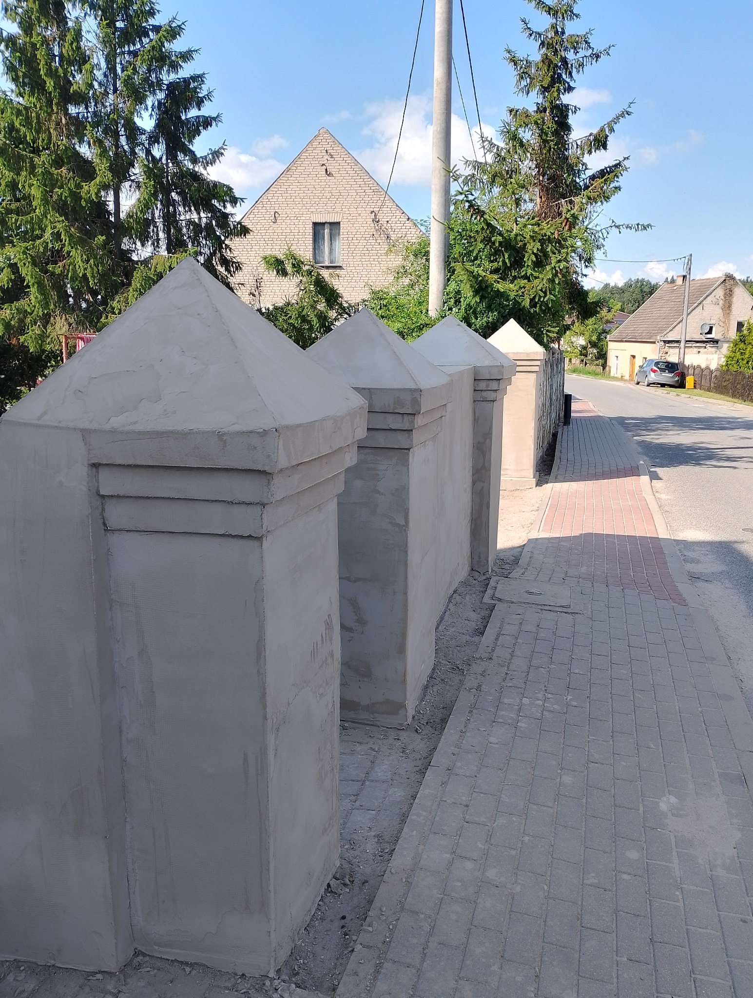Świeżo otynkowane słupki ogrodzeniowe z piramidalnymi zwieńczeniami, widoczne z boku na tle ulicy i domów. Detal wykończenia zewnętrznego w jasnym świetle.