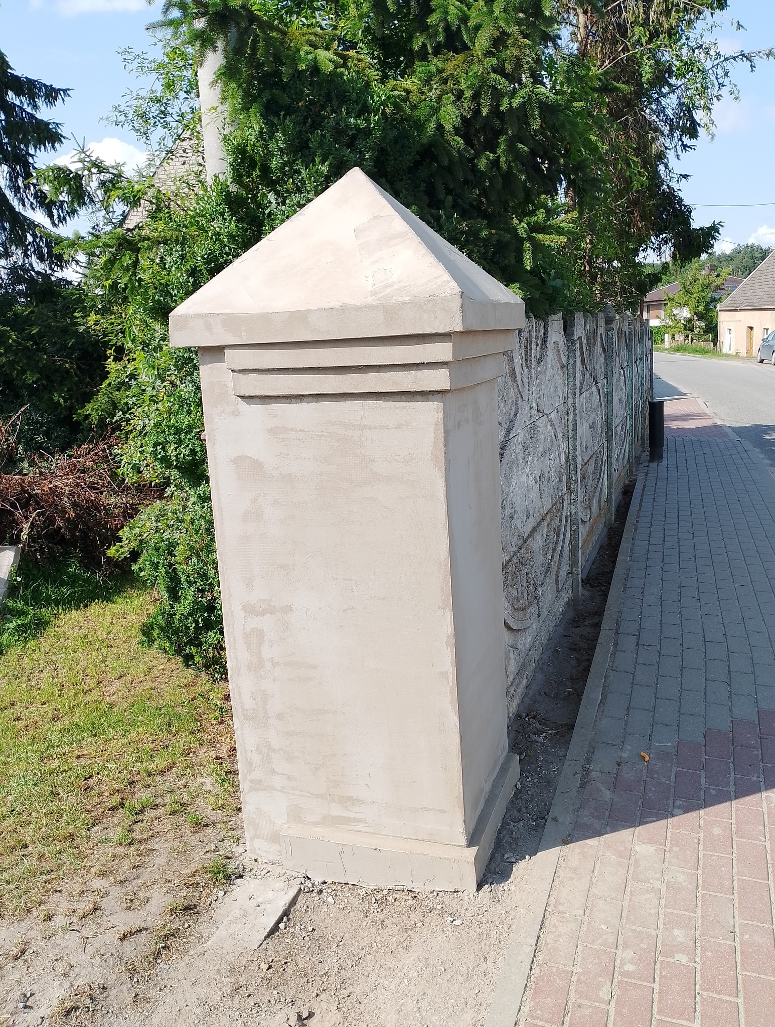 Wykończony fragment ogrodzenia z gładzią i dekoracyjnym wzorem, obok chodnik z kostki. Widoczne krzewy i drzewa w tle. Słoneczny dzień.