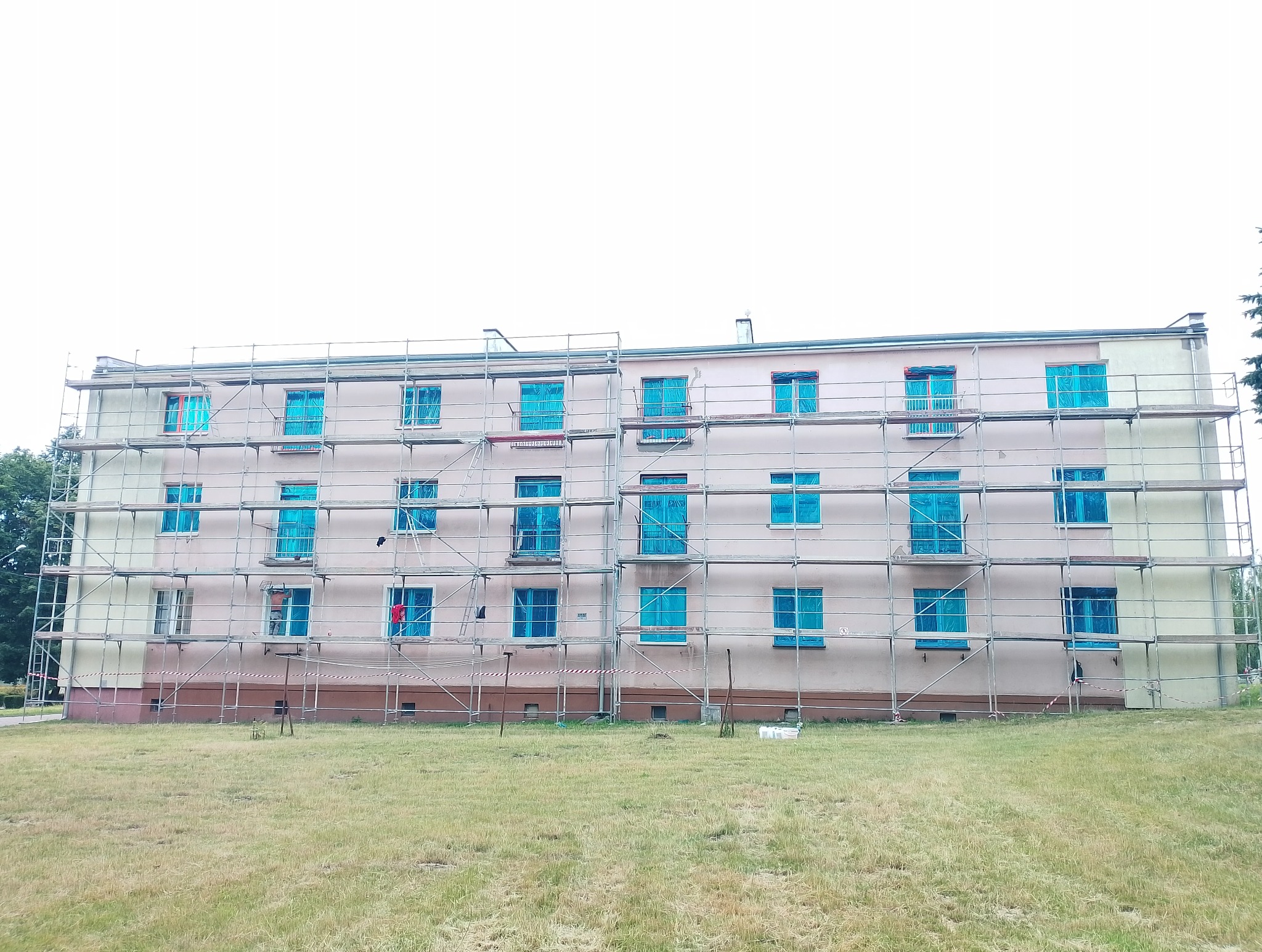 Budynek w trakcie malowania elewacji, zabezpieczone okna folią, rusztowania. Widoczny fragment pomalowanej ściany.