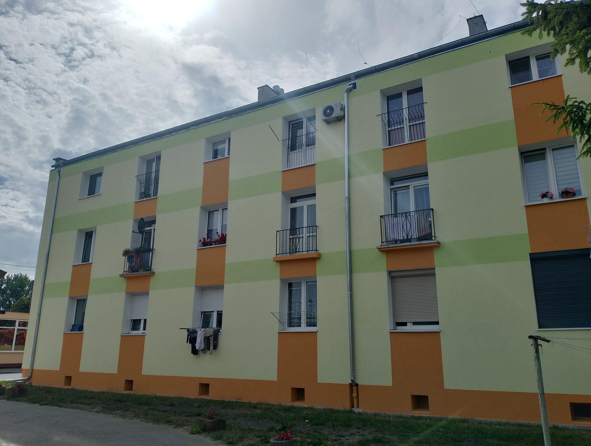 Wielopiętrowy budynek po renowacji, z balkonami i oknami. Elewacja w odcieniach zieleni i pomarańczy, z widocznymi detalami architektonicznymi. Na balkonie suszące się pranie.